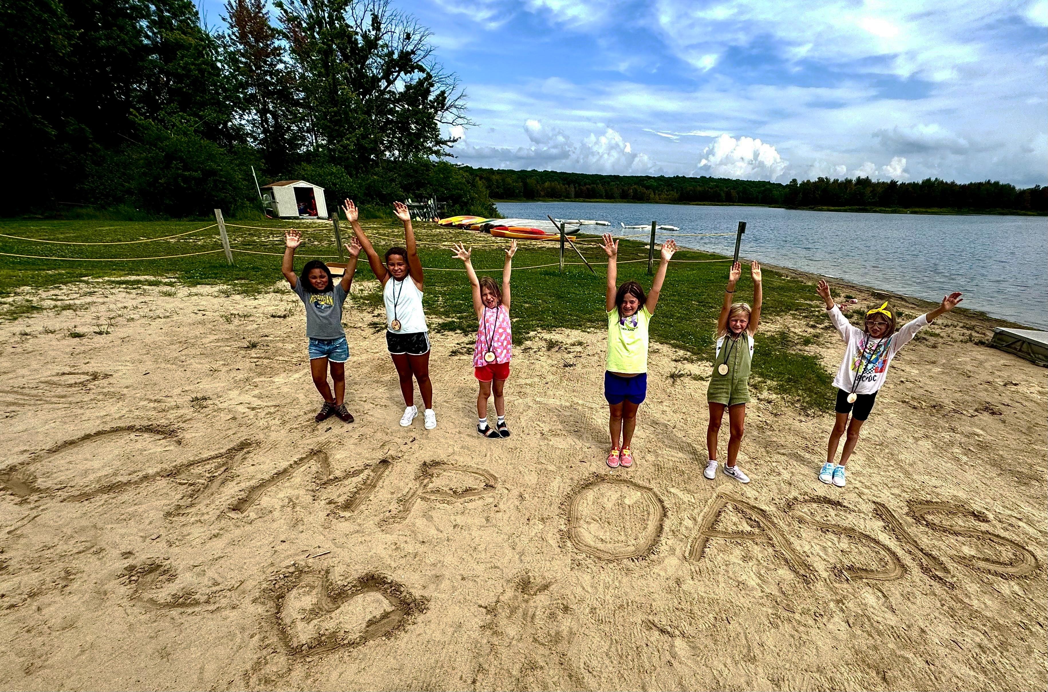 Campers stand with raised arms next to the words "Camp Oasis" drawn into sand