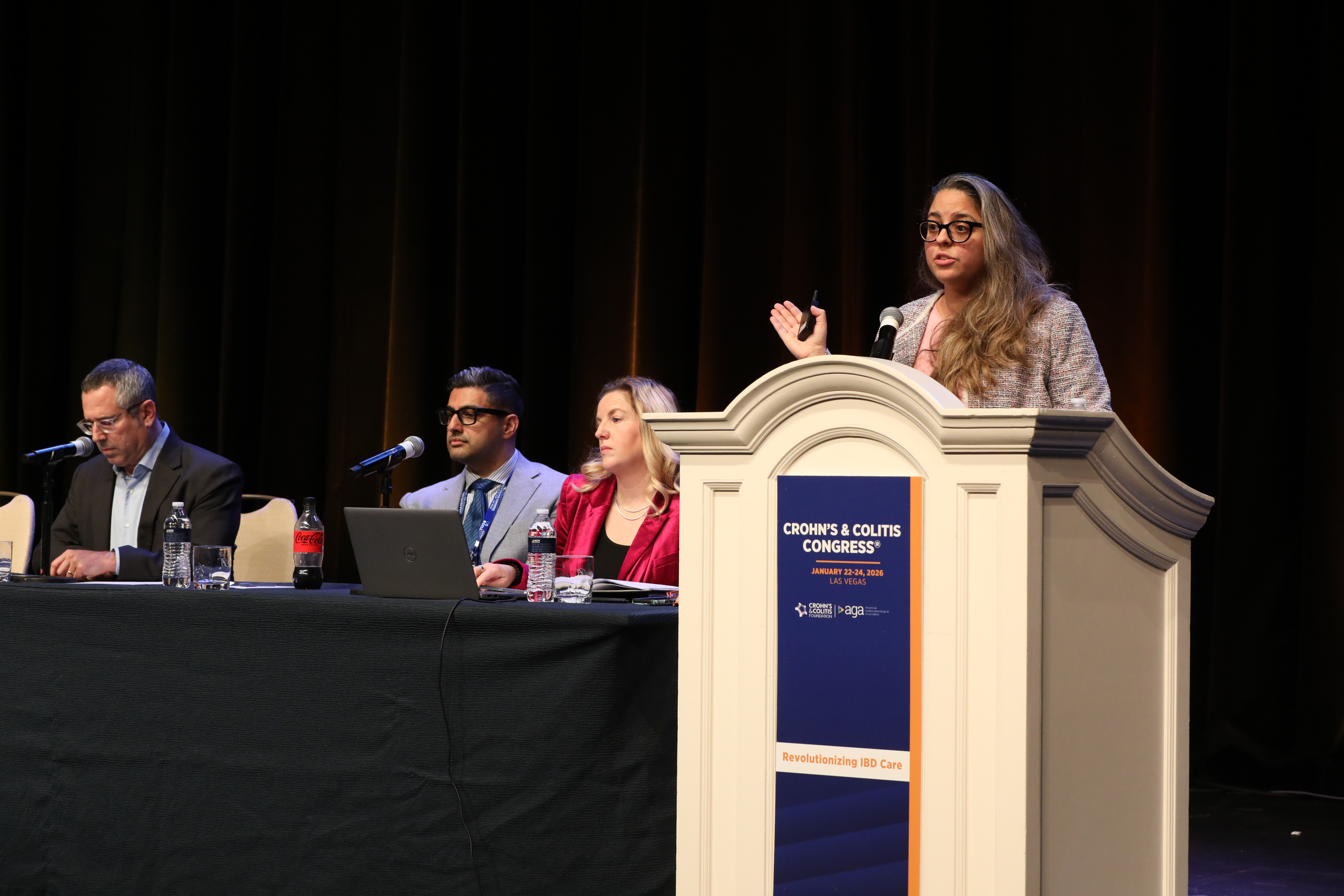 A woman speaks at a podium labeled "Crohn's & Colitis Congress" with three seated panelists beside her. The setting is professional and formal.