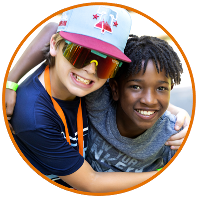 Two boys smiling and hugging, showing friendship and joy. One wears a cap and sunglasses, the other has a gray t-shirt.
