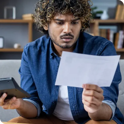 A man with curly hair sits on a couch, looking confused as he examines a piece of paper in one hand and holds a tablet in the other, with bookshelves in the background.