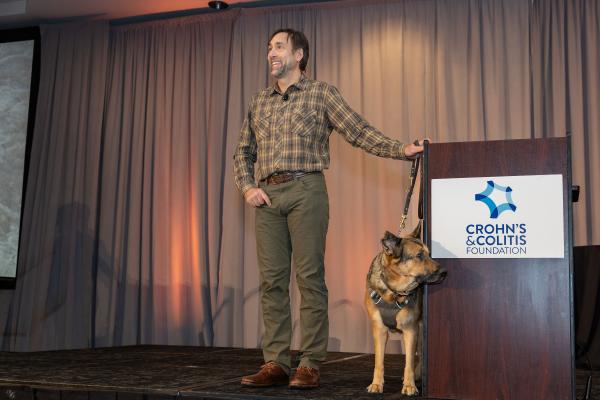 Man in plaid shirt and green pants smiles on stage beside podium. A German Shepherd sits calmly next to him. Behind, a curtain with warm lighting.