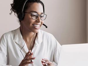 Smiling woman wearing a headset and glasses, seated at a desk with a laptop, appearing engaged and professional in a bright, minimalist room.