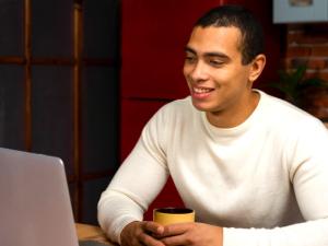 Man in a white sweater happily looks at a laptop while holding a yellow mug. He sits at a wooden table, conveying a relaxed atmosphere.