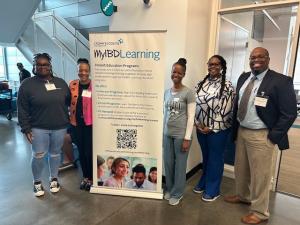 A group of five smiling people stand beside a "MyIBDLearning" banner detailing patient education programs. The setting is a modern indoor space.