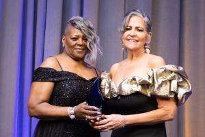 Two women in elegant, off-the-shoulder gowns stand smiling at an award ceremony. One holds a blue glass award. The mood is celebratory.
